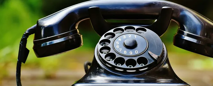 Close-up of a classic black rotary phone outdoors with a blurred green background.