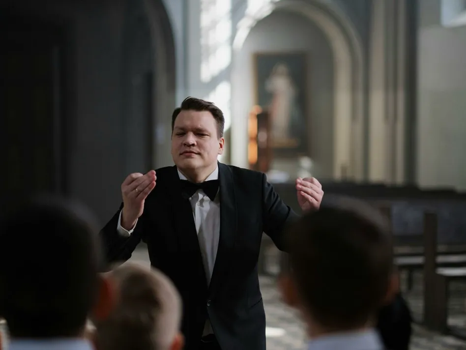 A male conductor leads a boys' choir rehearsal in a serene church setting.