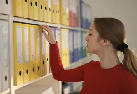 Side view of contemplating female assistant in casual style standing near shelves and choosing file with documents
