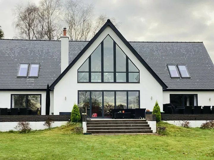 A modern two-story house with a steeply pitched roof and large triangular central windows. The home has white walls with dark roof shingles and black window frames. Two wings extend from the central structure, each with skylight windows