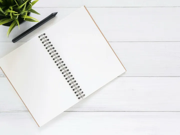 Open spiral notebook on white desk with black pen and green plant. Ideal for planning.