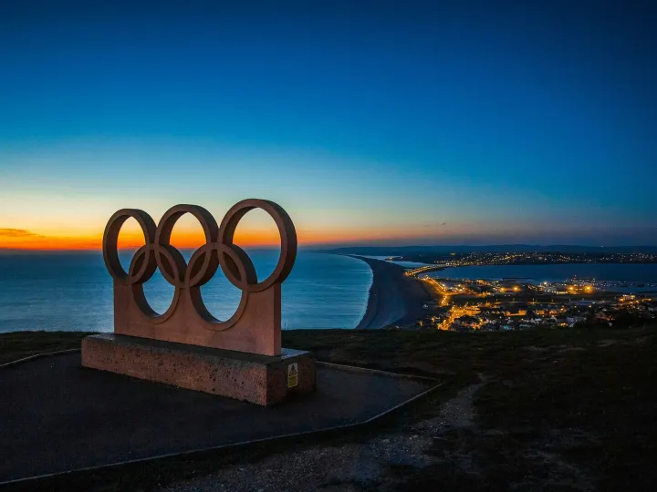 Scenic sunset view of the Olympic rings monument by the sea with city lights in the distance.
