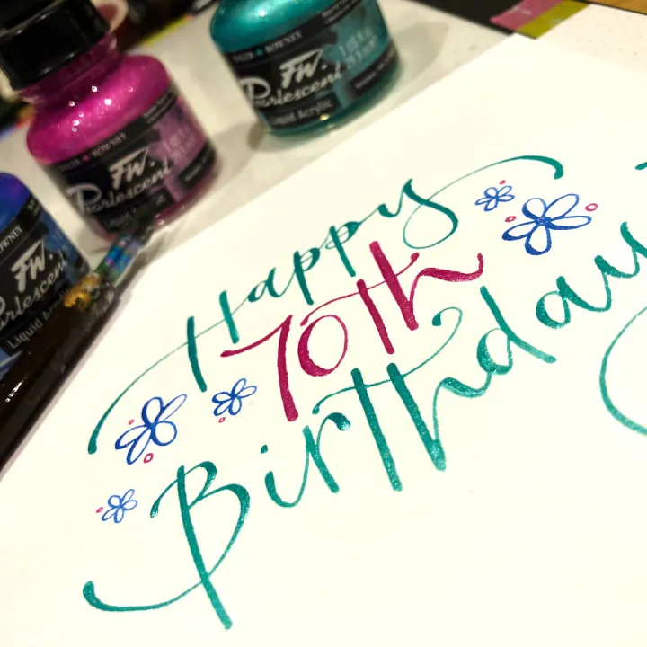 Happy 70th birthday card lettered in blue and pink beside pots of ink