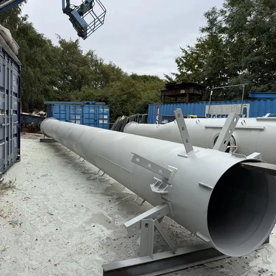 Long primed grey steel flue section resting on steel stands in an outdoor yard, with a second section behind