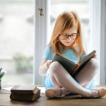 Adorable young girl wearing glasses, reading a book indoors by a window. Captures innocence and focus.