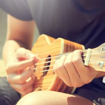 Close-up of a man's hands strumming a ukulele outdoors on a sunny day.