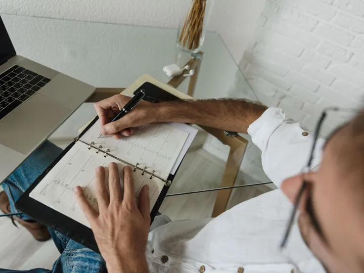 A man writing in a diary planner beside a laptop