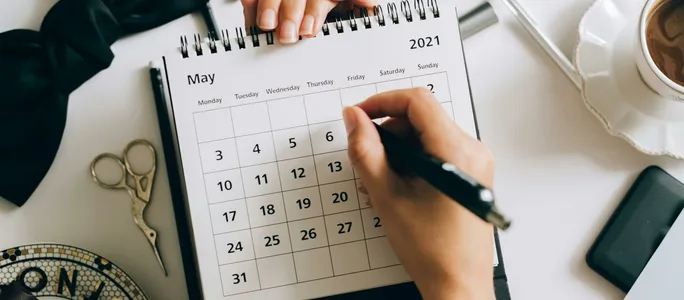 Top view of hands using a planner with coffee and accessories, focused on May 2021 calendar.