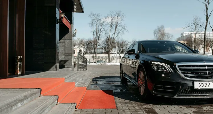 A luxury black sedan parked on a red carpet driveway outside a modern building.