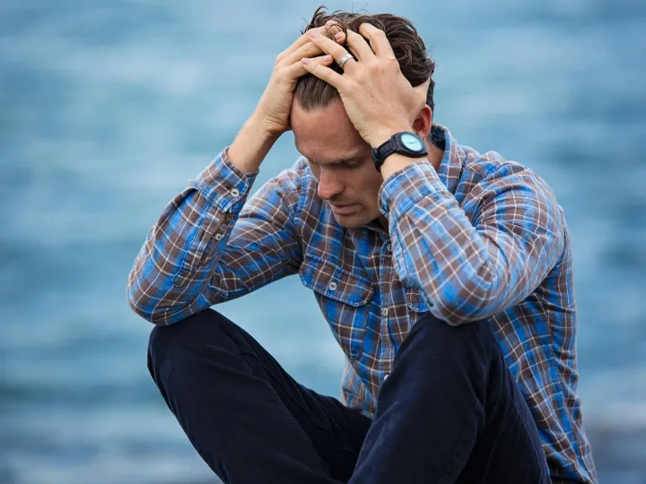 A man in a plaid shirt sits by the water looking