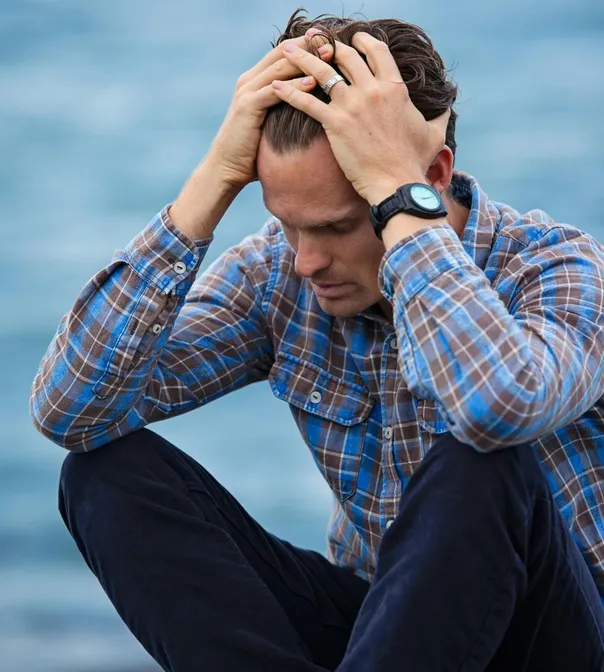 A man in a plaid shirt sits by the water looking