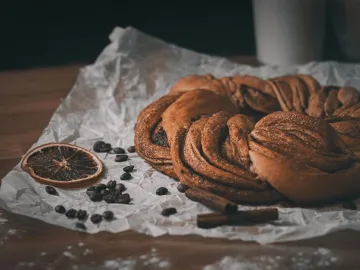 Close-up of a delicious cinnamon pastry on paper, accompanied by coffee beans and a dried orange slice.