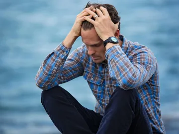 A man in a plaid shirt sits by the water looking
