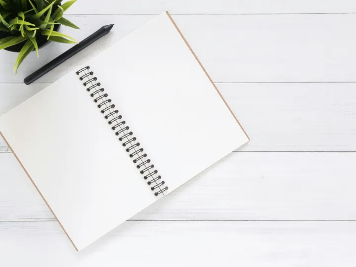 Open spiral notebook on white desk with black pen and green plant. Ideal for planning.