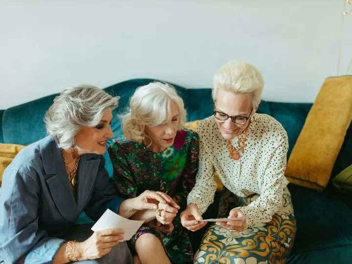 Three senior women joyfully reminiscing over old photos on a cozy couch.