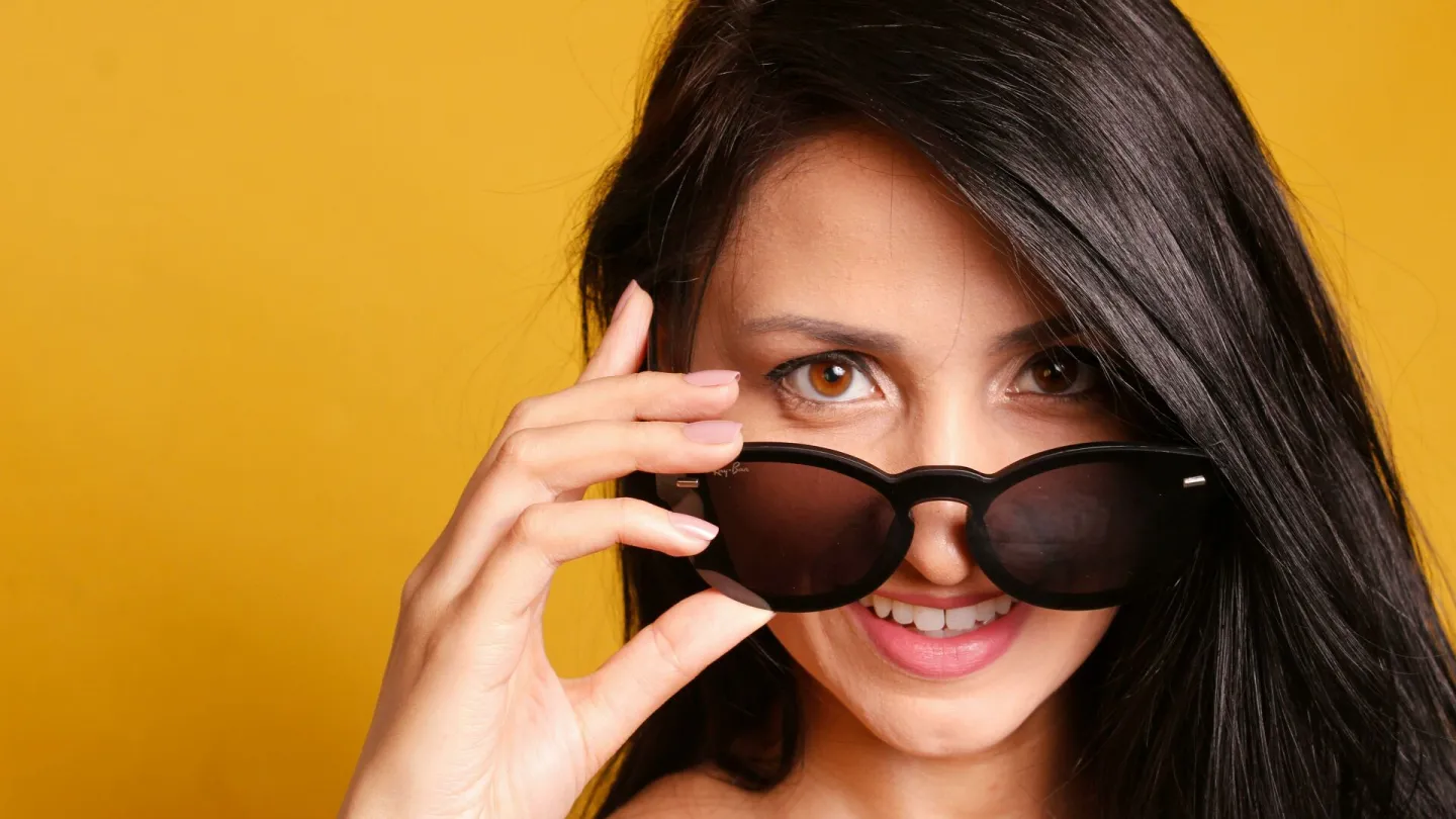 Portrait of a smiling woman with sunglasses posing against a vibrant yellow background, exuding style and confidence.